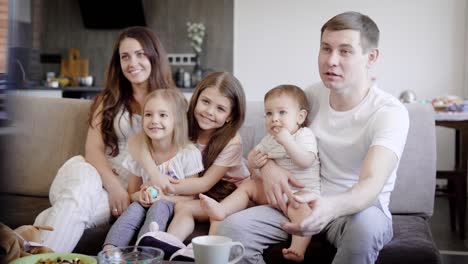 happy family couple with three little daughters are sitting on a couch in a living room and watching tv in daytime