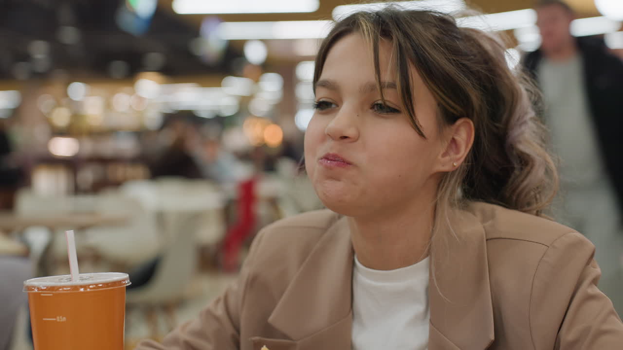 어린 학생이 패스트푸드를 먹고 있어요. 밝은 옷을 입은 십 대 청소년이 빠른 식사를 열정적으로 즐기고 있으며, 십 대 소년은 바쁜 식당 환경에서 햄버거와 소다를 열심히 먹고 있어요.