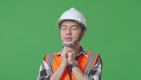 close up of asian male engineer with safety helmet pray for something while standing in the green screen background studio