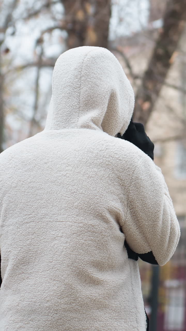 겨울 재을 입은 사람이 고 전화로 대화하는 뒷면으로, 흐릿한 배경에는 건조한 나무와 건물이 있으며, 도시 환경에서 평화로운 겨울 장면을 불러일으킨다.