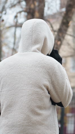 vista trasera de una persona con chaqueta de invierno caminando y hablando por teléfono con un fondo borroso con árboles secos y edificios, evocando una escena de invierno pacífica en un entorno urbano