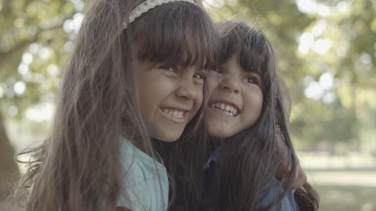 Primer plano de retrato de niñas latinas abrazándose, sonriendo y dándose un beso en el parque.
