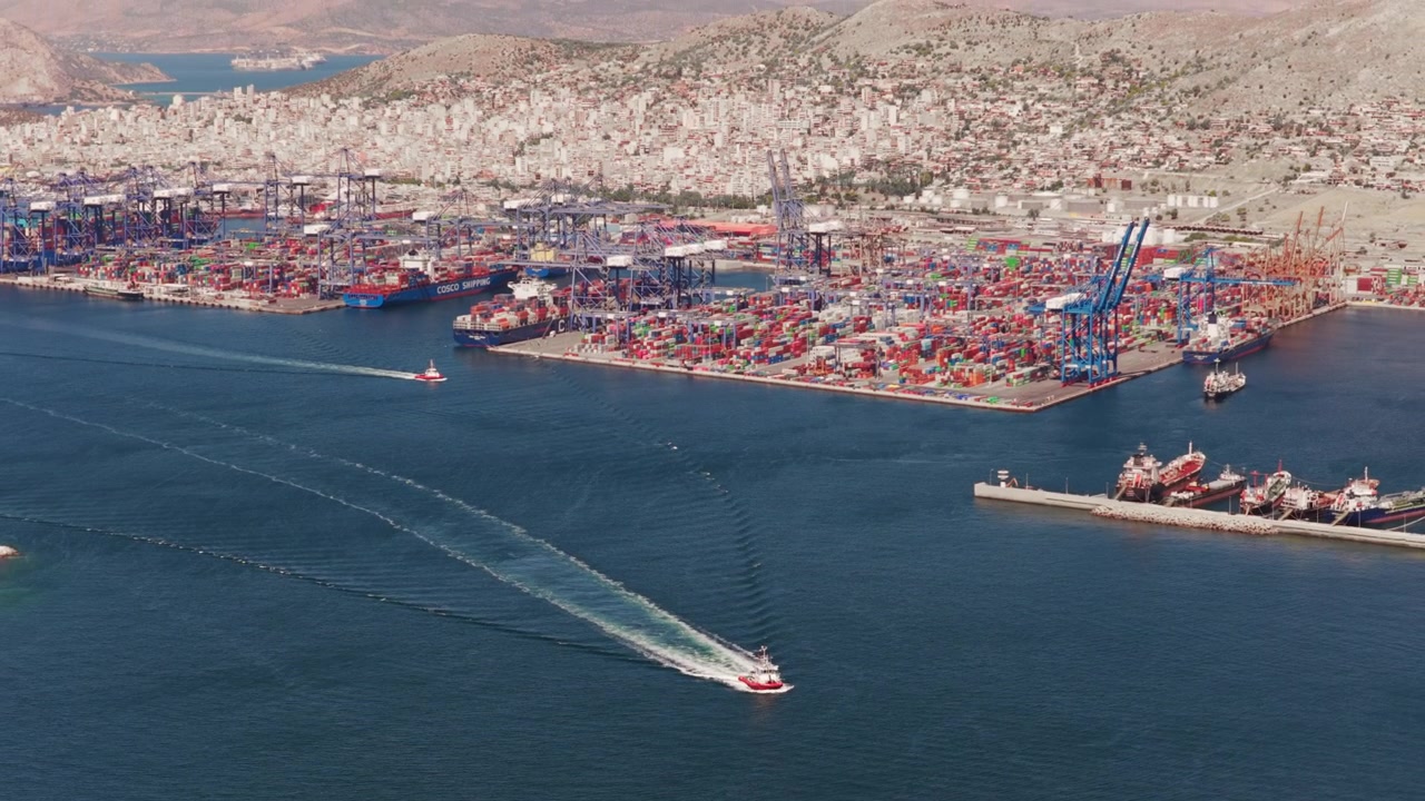 Expansive aerial view of the Piraeus container port in Greece. Showing a pilot boat navigating the harbor with massive cranes. Stacked cargo containers. And the sprawling cityscape in the background
