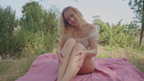 an attractive woman sits on a picnic blanket in a sunny forest and strokes her legs in slow motion while smiling at the camera