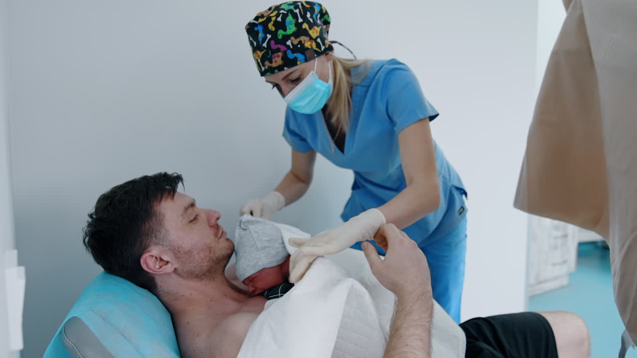 Tiny newborn is put on the bare chest of a father. Nurse is helping to cover the baby with sheets and blankets.