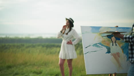 a painter, partially visible, is creating a portrait of a woman who stands poised in a white dress and black hat. the setting is a tranquil grassy landscape