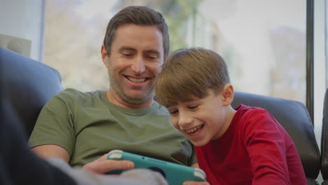 father with son wearing pyjamas sitting on sofa at home playing computer game on handheld device - shot in slow motion