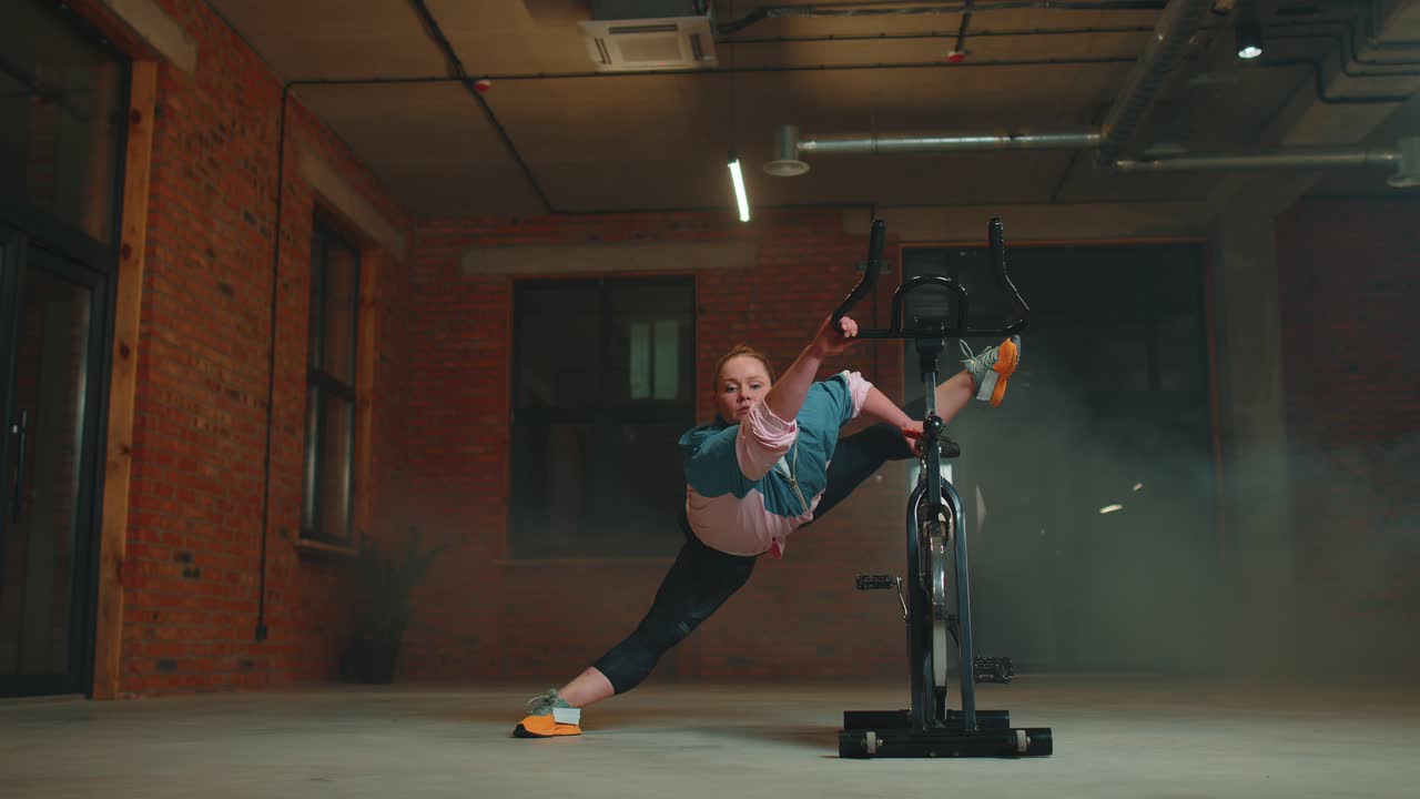 Athletic woman makes twine training routine on spinning stationary bike in gym stretching exercises