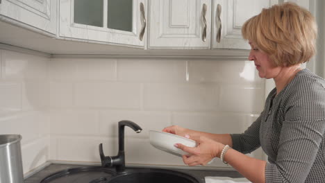 woman removing dirt from white plate while placing it carefully under kitchen cabinet with a modern black sink and faucet in background, performing dishwashing task in kitchen