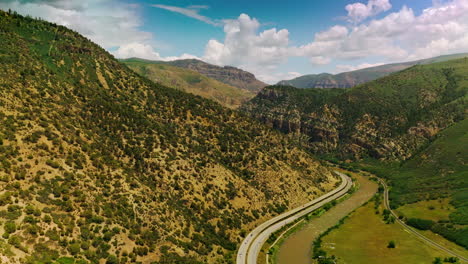 cars running along the road going around the green mount. sunny picturesque view of mountains at the backdrop of cloudy blue sky.