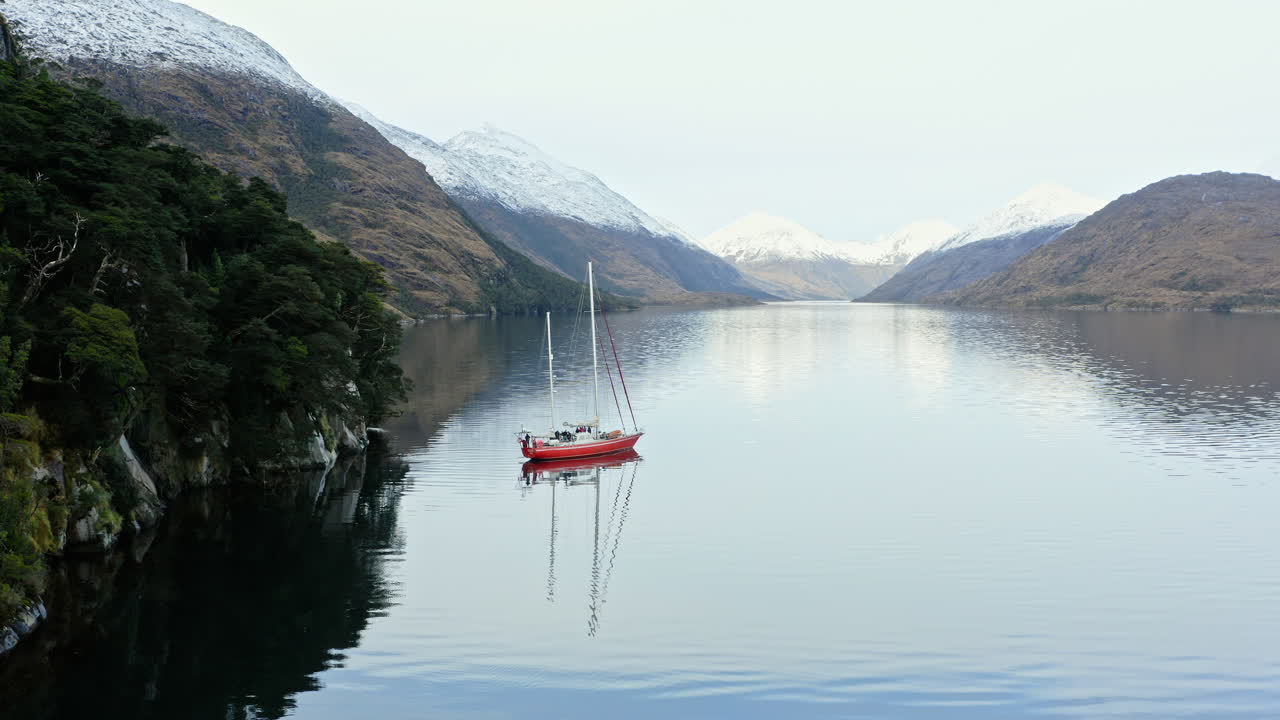 Sailboat travels between narrow fjords in Beagle Channel with high cliffs and smooth water, establishing descend aerial