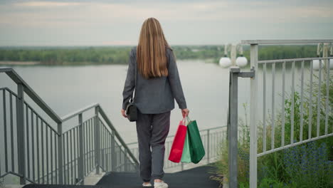 back view of lady carrying colorful shopping bags walking down stairs with two people ahead, background featuring river, greenery, and a lamp post along the staircase railings on a calm day