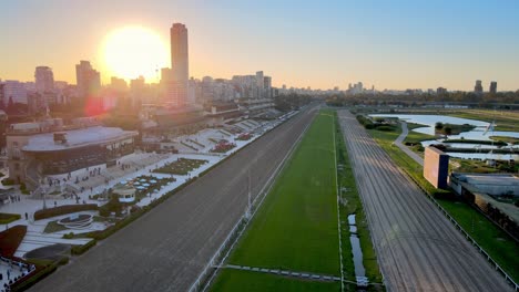 Langsamer-Luftwagen-In-Aufnahme-Entlang-Der-Pferderennbahn-Auf-Der-Berühmten-Rennbahn-Hipodromo-Argentino-De-Palermo-In-Der-Hauptstadt-Buenos-Aires,-Argentinien,-Mit-Stadtbild-Und-Sonnenuntergang-Im-Hintergrund