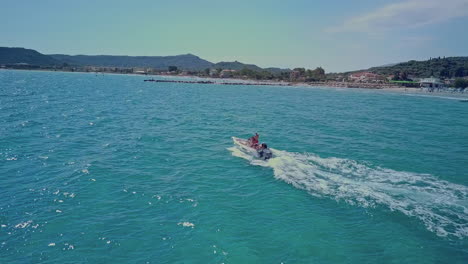 Aerial-following-small-boat-on-blue-water-off-shoreline-of-Greek-island