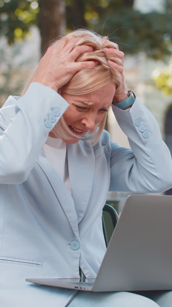 Upset mature caucasian businesswoman reading bad news on laptop sitting on bench on city street