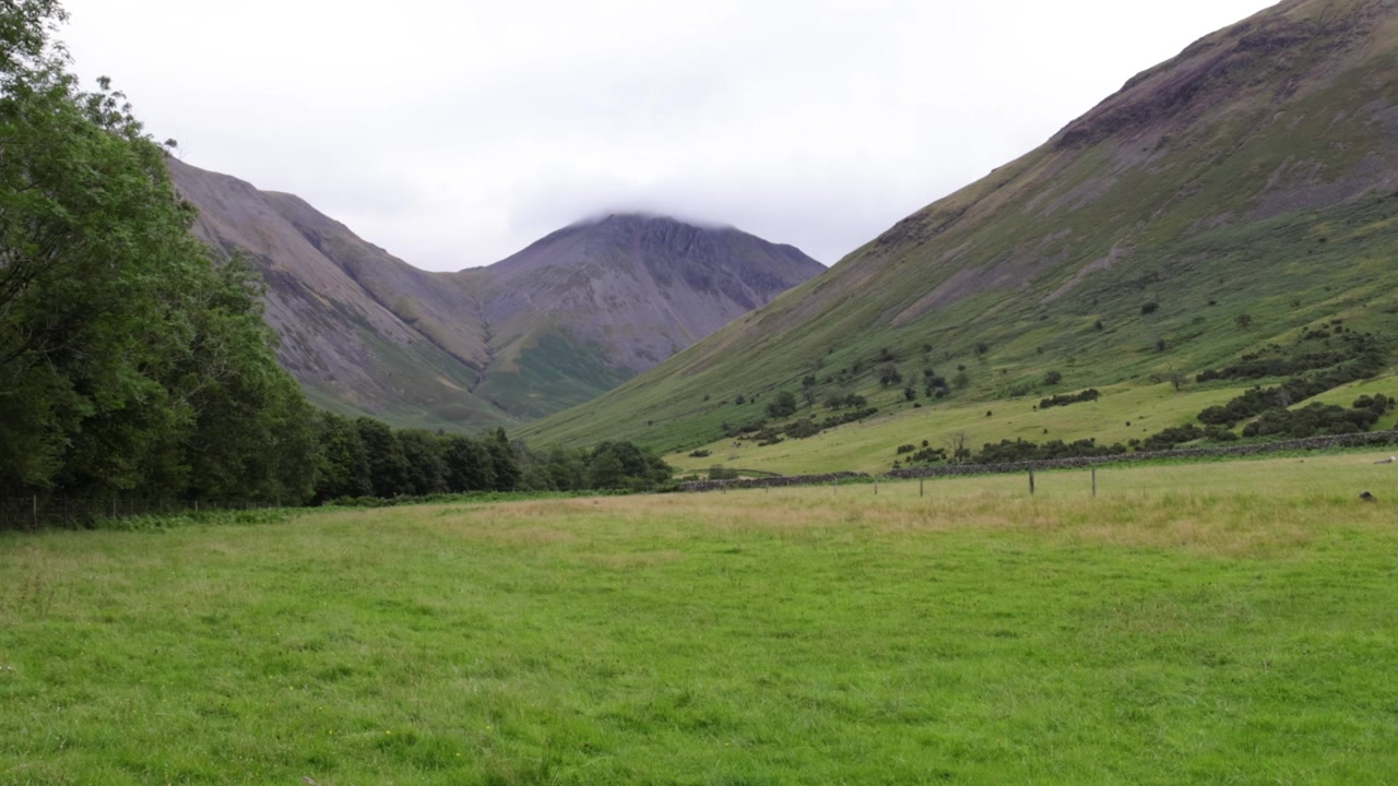 Il monte Great Gable si erge sopra il prato della valle di Seascale nel Lake District, Cumbria, Inghilterra. Ampia inquadratura statica di pascoli verdi, colline e nuvole basse sotto un cielo grigio
