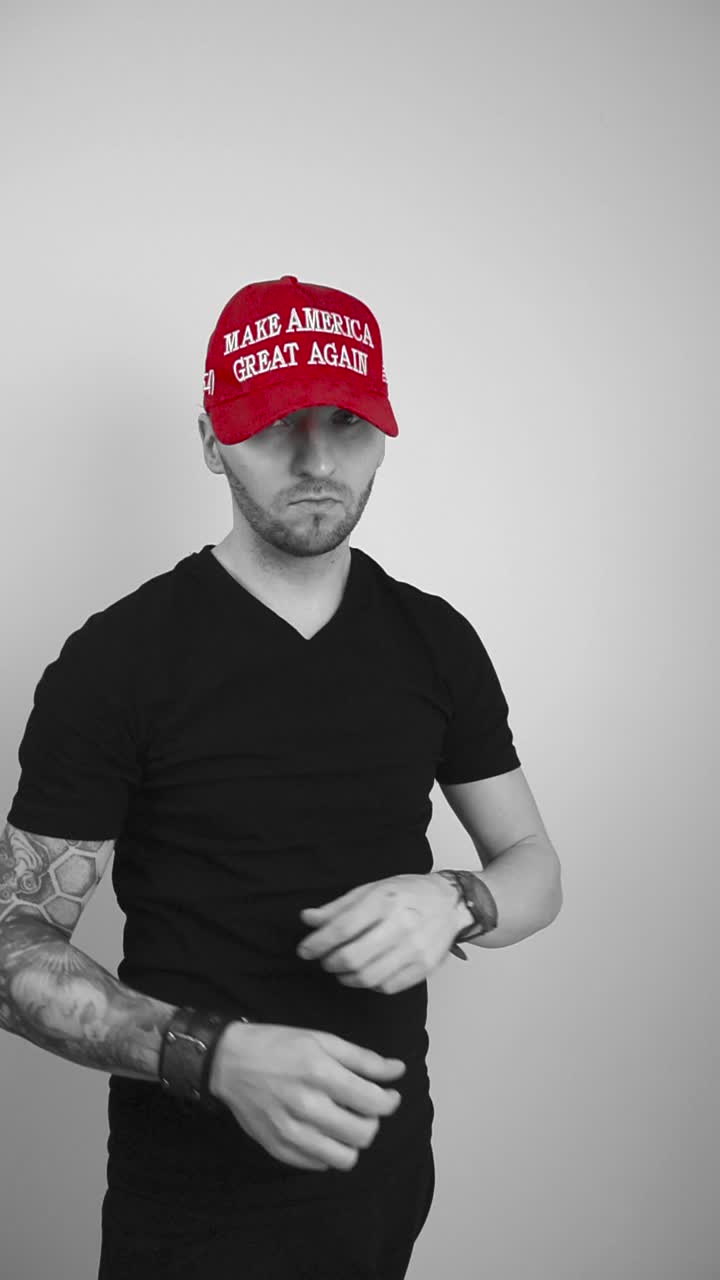 Grayscale footage of a man with full sleeve tattoo, short hair and beard putting on a red vibrant colored MAGA Make America Great Again hat, looking forward, crossing his arms and nodding happily.