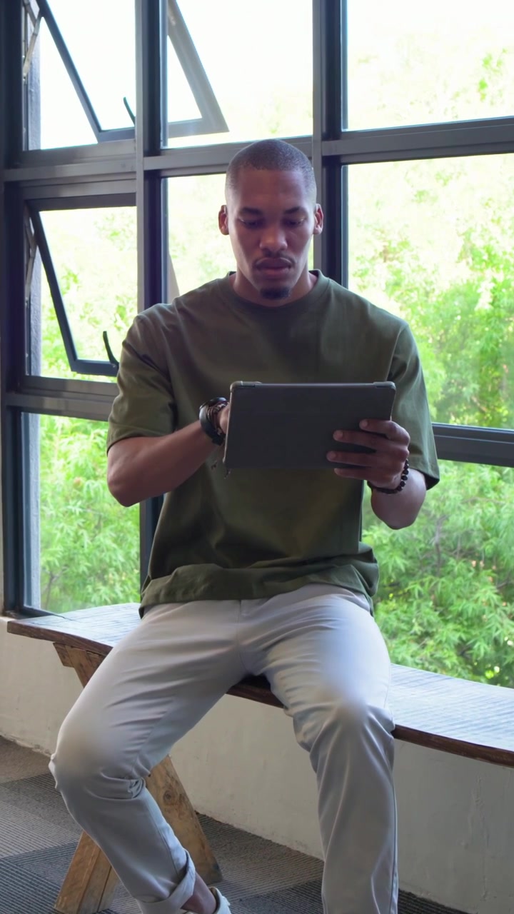 Vertical video: Pausing, tapping man in olive shirt using tablet on bench by window, for work