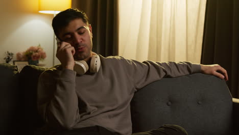 Young-Man-Wearing-Wireless-Headphones-Spending-Evening-At-Home-Sitting-On-Sofa-Talking-On-Mobile-Phone-2