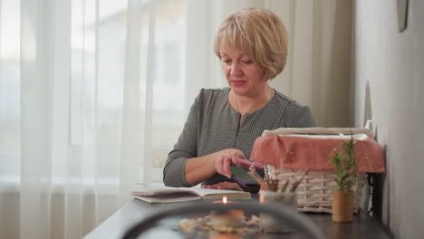 woman seated beside chair operating phone and opening new page of notebook with basket, candle and study items on desk in cozy room filled with natural light streaming through sheer curtains