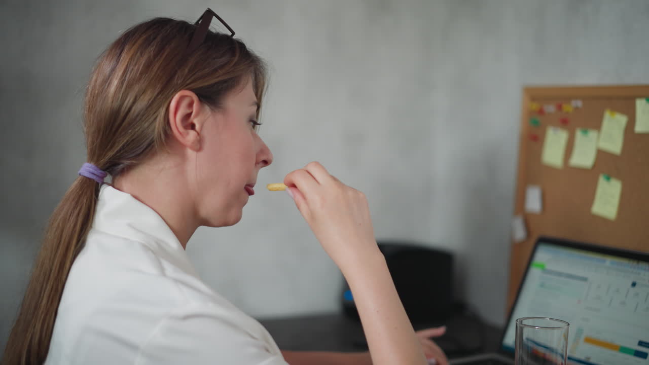 Freelancer woman with hair tied back wearing white shirt multitasking at workspace eating fries while working on laptop surrounded by organized corkboard and documents in cozy modern home office