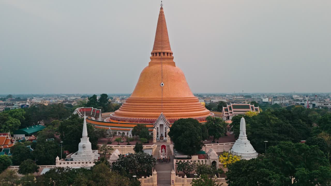 La prospettiva aerea inizia vicino all'enorme stupa arancione, poi si allontana lentamente rivelando i grandi gradini d'ingresso bianchi circondati da lussureggianti alberi verdi sotto cieli mattutini nuvolosi e foschi.
