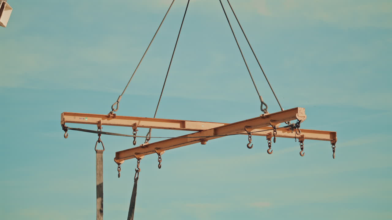 A yellow metal lifting beam with hooks, suspended by cables against a clear sky