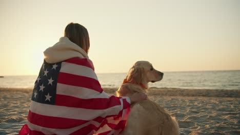 close-up shot a blonde girl sits on a sunny beach with her dog wrapped in an american flag on a sunny beach in the morning