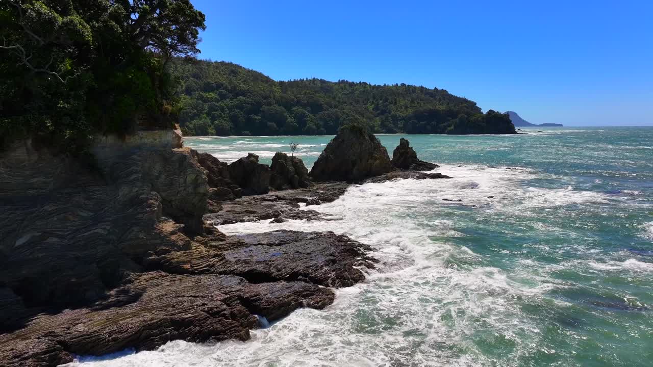 Moody Aerial beech Unspoilt jagged rocky coastline—turquoise waves crash against cliffs, windswept trees cling to edges, sunlight flickers across the wild, untouched shore. Drone New Zealand native