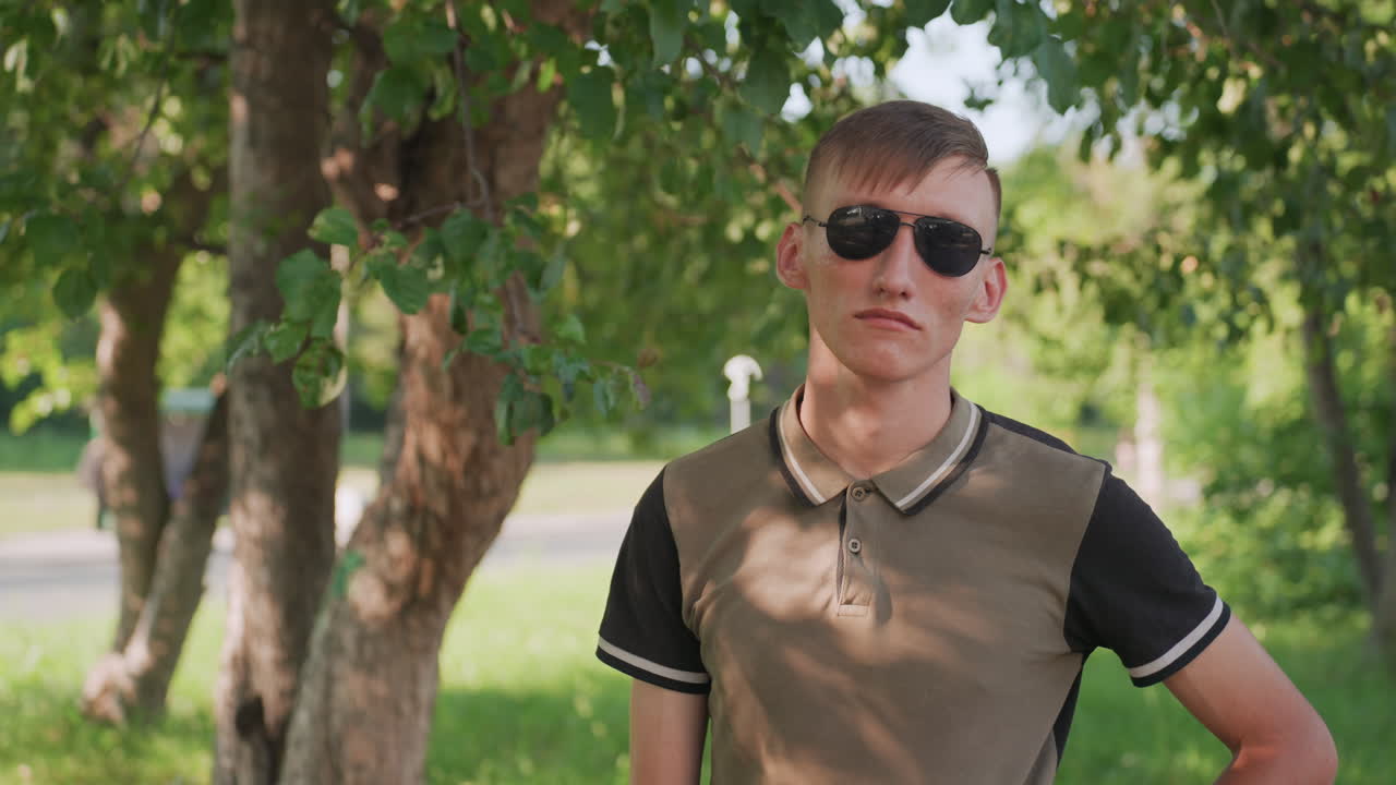 Man In Sunglasses Stands Proudly, Young Individual Displays Relaxed Selfassurance Outdoors, Young Man Stands Confidently With Hands On Hips Amidst Sunlit Greenery Wearing Casual Attire