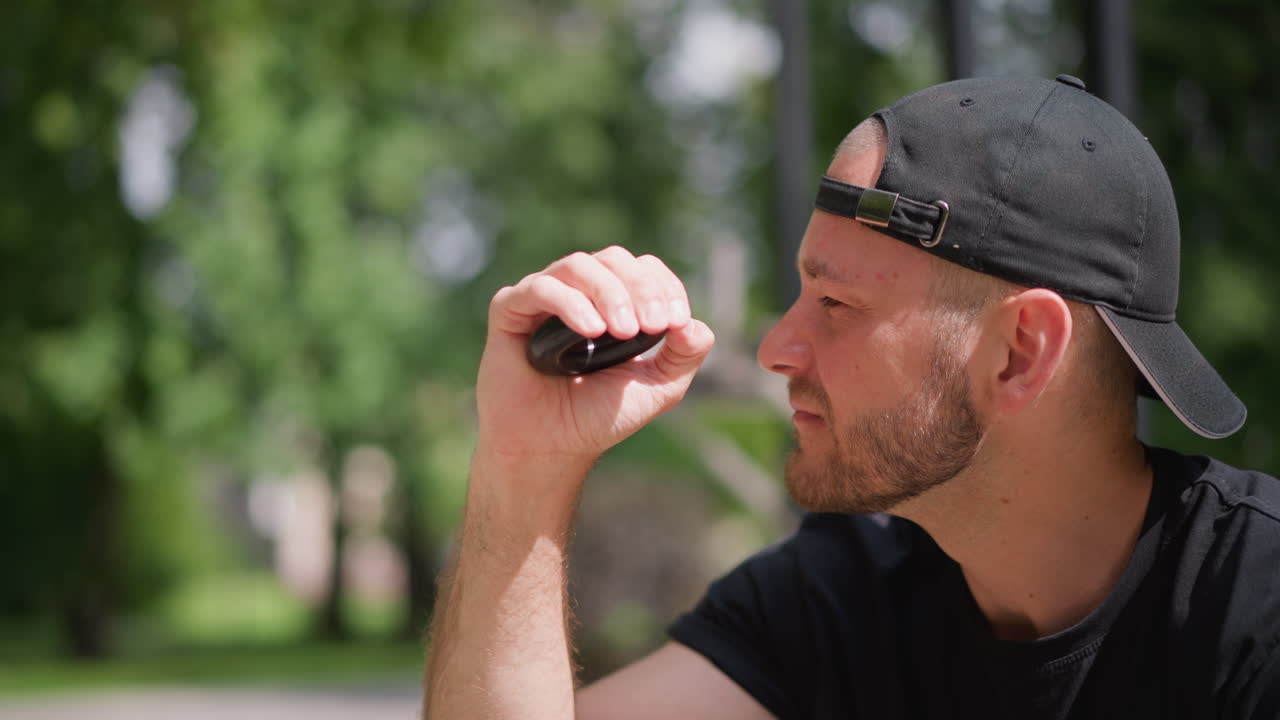 작은 장치를 들고 미소 짓는 남자, 휴대용 기기를 들고 앉아 있는 남자의 자연스러운 야외 장면, 편안한 표정과 뒤로 쓴 모자, 부드러운 자연광과 배경의 건설 발판이 연출되었어요.