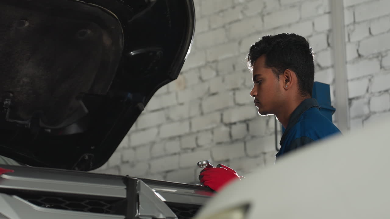 파란 유니폼과 은 장갑을 입은 자동차 수리업자는 오토 워크에서 열려있는 자동차 엔진을 검사하는 동안 스파이너를 들고, 배경에서 밝은 빛이 빛나고, 집중된 메카닉에 극적인 효과를 만니다.