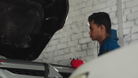 car repairer wearing blue uniform and red gloves holds spanner while inspecting open car engine in auto workshop, bright light shines from background, creating a dramatic effect on focused mechanic