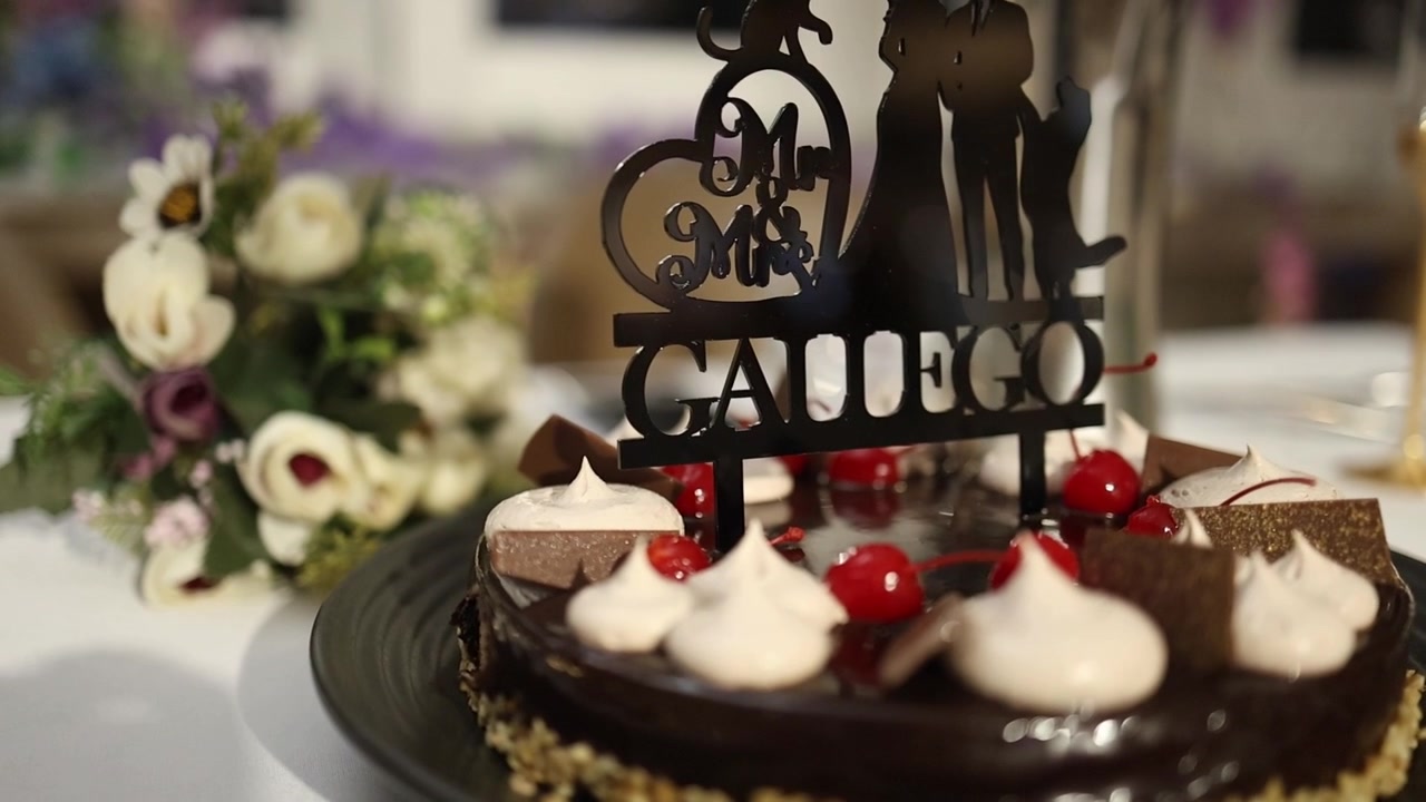 Cận cảnh chi tiết bánh cưới sô cô la với quả anh đào, kem tươi và phụ kiện trang trí cá nhân hóa, cùng với một bó hoa, đặt trên bàn tiệc dưới ánh sáng tự nhiên dịu nhẹ