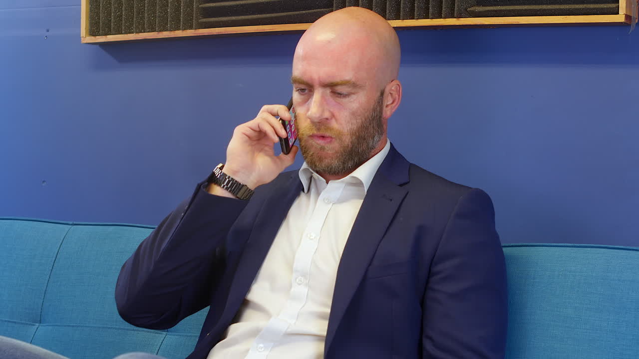 Uomo d'affari in completo che ascolta qualcuno al suo cellulare mentre è seduto su un divano nell'ufficio domestico