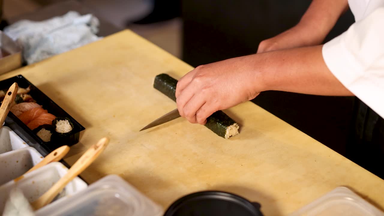 Chef professionista taglia un rotolo di maki alle alghe su un tagliere di legno nella calda illuminazione della cucina di un ristorante