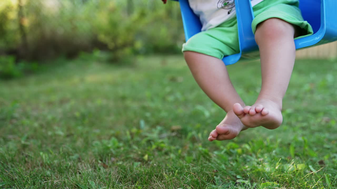 Little feet of a baby sitting on a swing. Barefoot child having fun outdoors. Close up. Blurred backdrop.