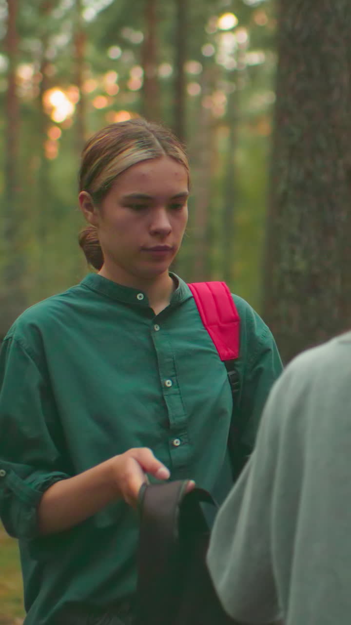 joven con camisa verde ayuda a su amiga con pañuelo azul a ponerse la mochila en el bosque, la luz del sol se filtra cálidamente a través de los árboles, iluminando la escena mientras se miran en silencio