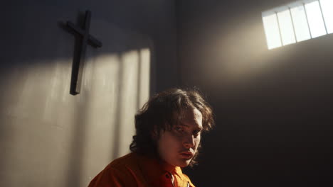 portrait of young man in prison cell sitting under wall cross