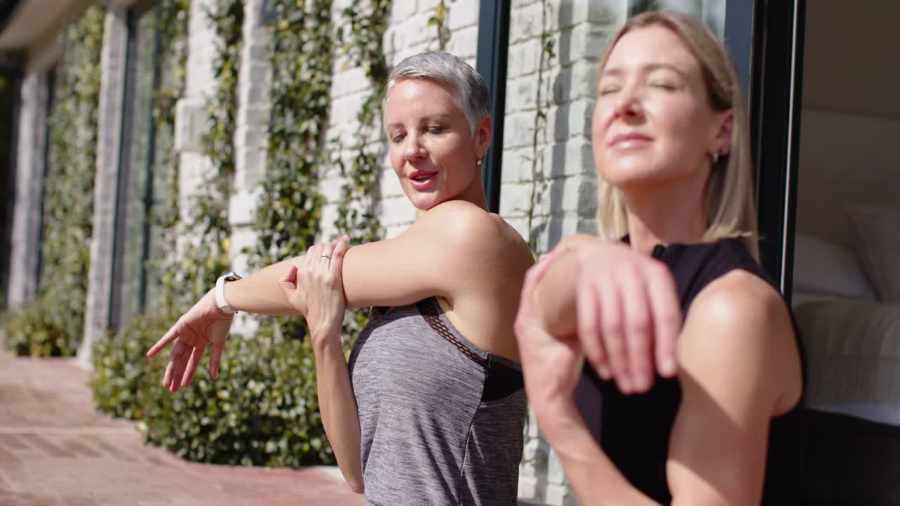 Partner donne che imitano l'istruttore sollevando il braccio destro con l'orologio e scaldando le spalle sul patio.