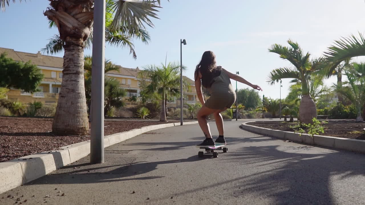 Embracing the surroundings, slow motion reveals a lovely young girl cruising on her longboard along the road near the beach and palm trees