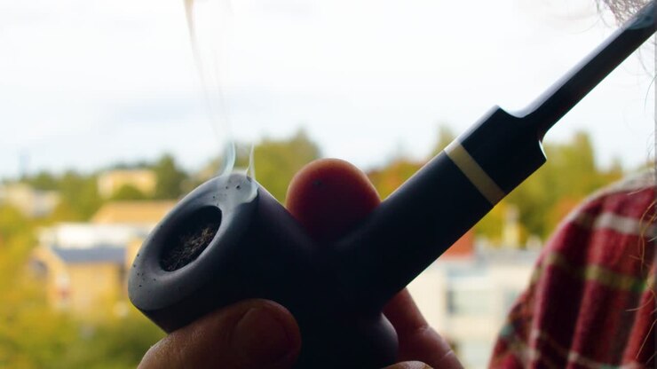 Caucasian male smoking holding a black vintage antique pipe producing thick smoke with quality western tobacco enjoying his day off on balcony at home on sunny day houses and nature in the background