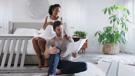 pareja interracial con una mujer embarazada mirando las instrucciones para construir una cuna para bebés de autoensamblaje