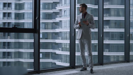 Young-businessman-searching-tablet-computer-standing-by-window-in-office-centre.