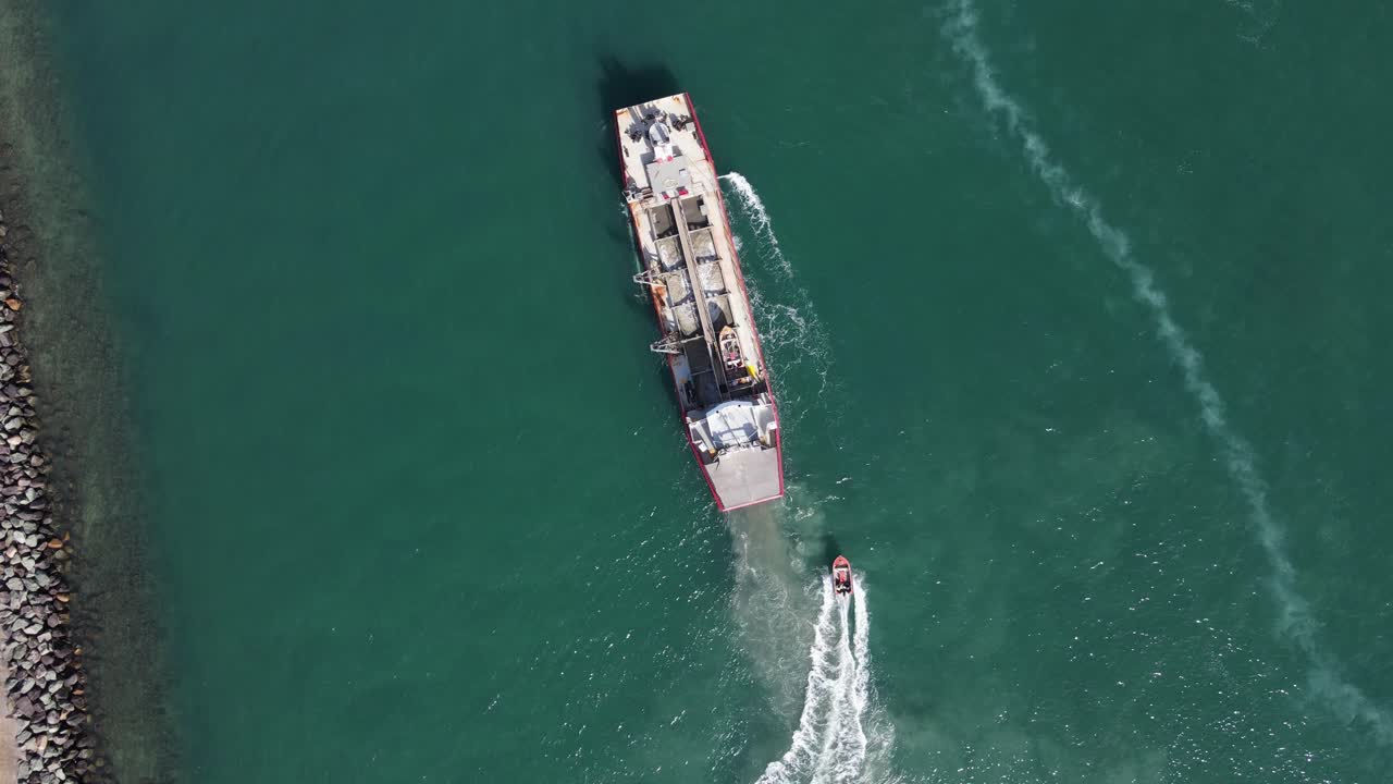 una gran draga industrial maniobra en el agua mientras un rápido barco piloto navega junto al barco.