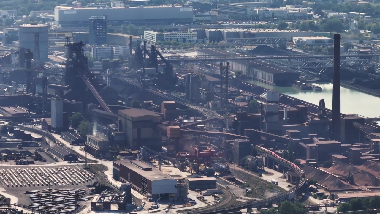 Large steel factory with smokestacks rising behind a dense green forest. The industrial landscape shows heavy manufacturing and production