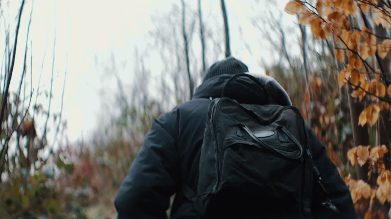 man with a black backpack hiking up a hill on a cloudy autumn day. swirley bokeh in 4K Sow Motion. fall landscape.