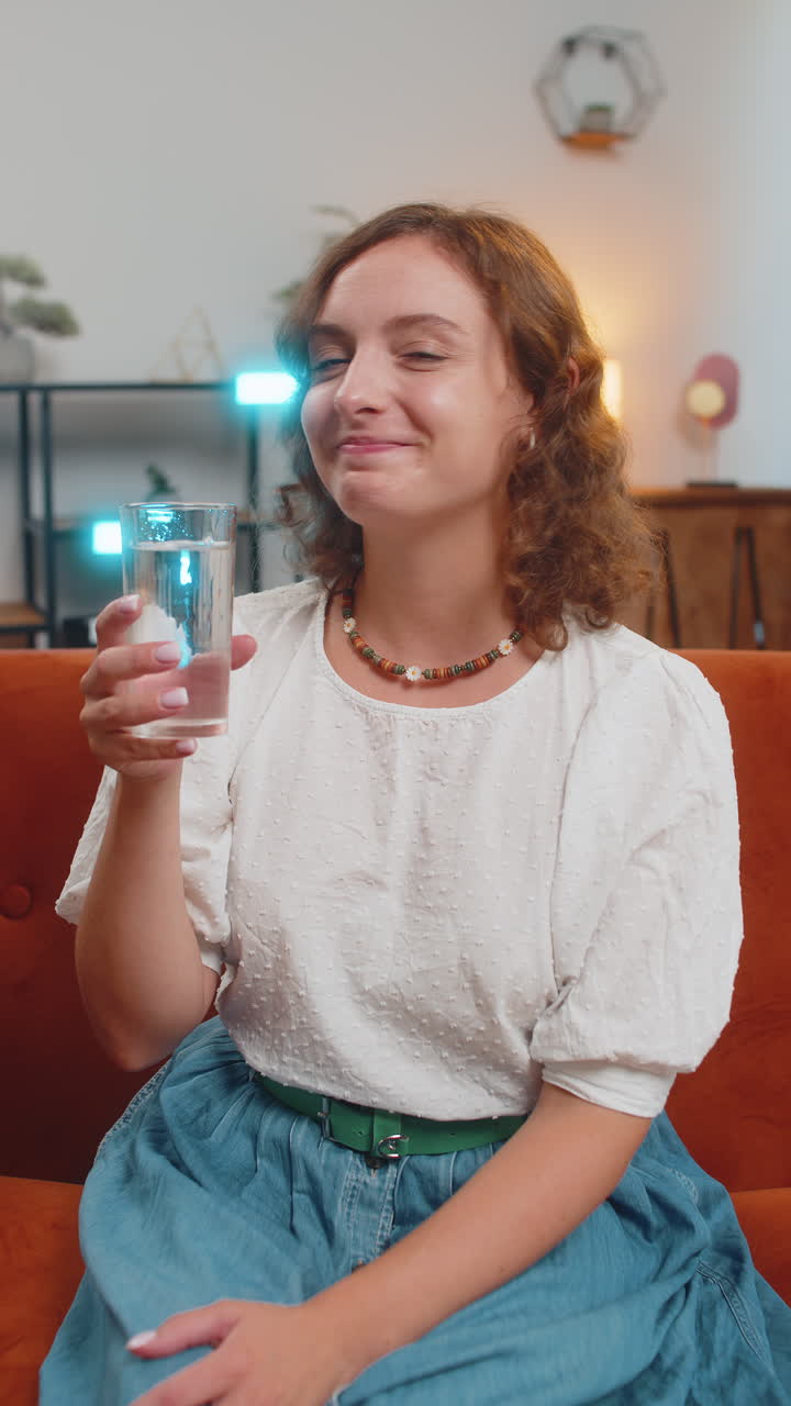 Happy woman sitting on sofa couch leading healthy lifestyle drinking a glass of pure water at home