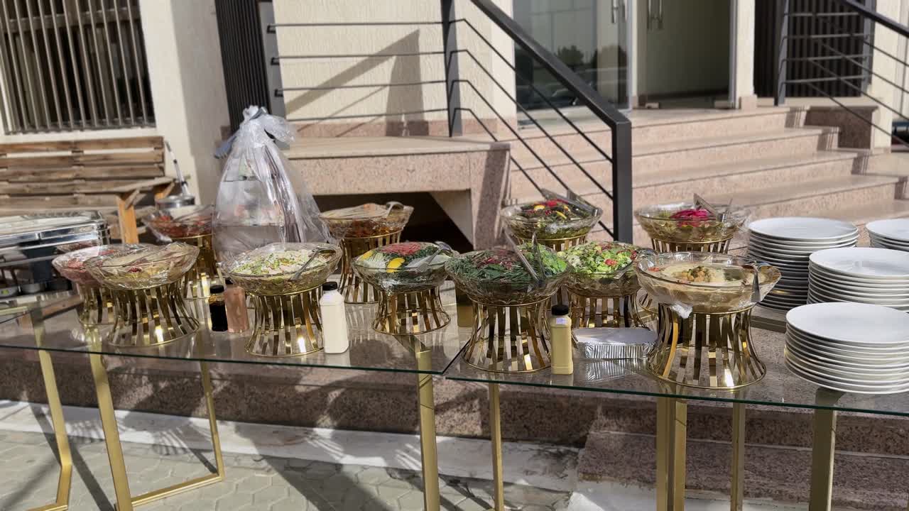 Buffet soleado al aire libre en un patio con calentadores y ensaladeras cubiertas en mesas de cristal, ideal para una recepción de boda o un evento de comida corporativa.