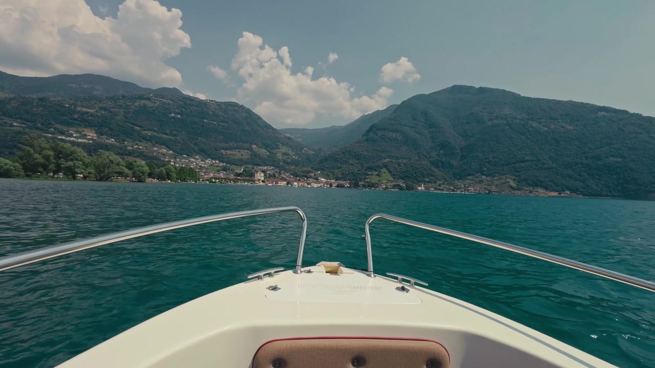 POV dalla prua di una barca che scivola su acque profonde e blu di un lago con maestose montagne davanti, catturando la sensazione di una lussuosa vacanza estiva sotto un cielo azzurro limpido
